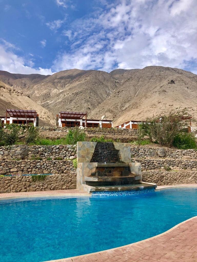 A swimming pool is surrounded by a stone wall, featuring a waterfall that flows gently into the clear blue water. In the backdrop, mountains rise majestically, and several wooden cabanas with shaded porches are visible along the hillside.