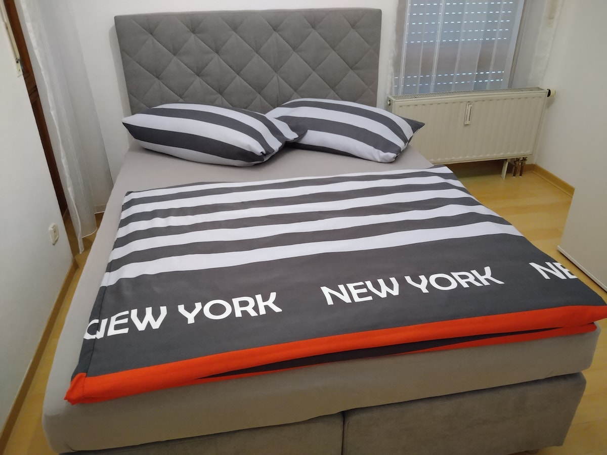 The sleeping area features a spacious bed with a upholstered headboard and coordinating striped bedding. Two pillows are placed atop the bed, complementing a grey and orange blanket. Natural light enters through a partially visible window with a fabric treatment.