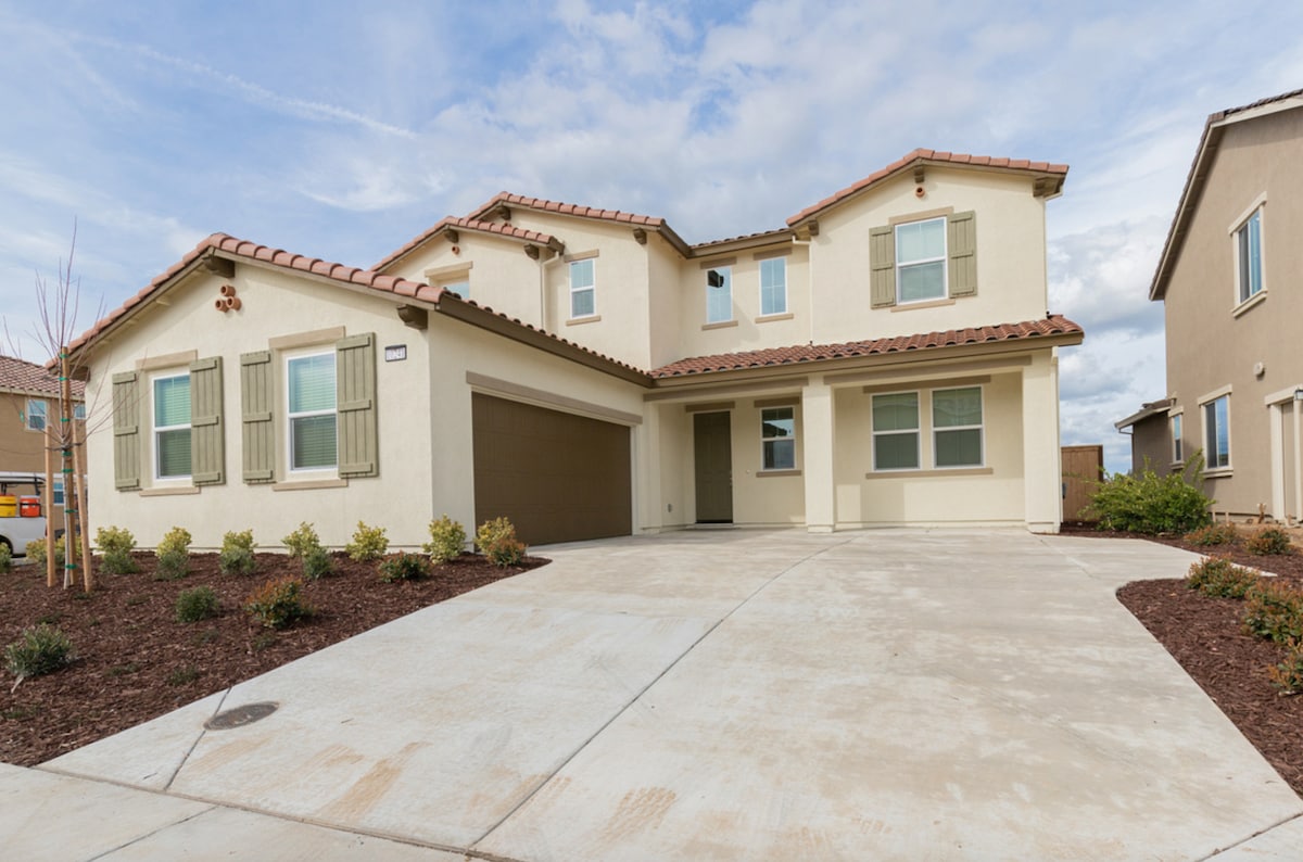 A two-story house features a warm beige exterior with brown roof tiles. The front porch is flanked by large windows with green shutters. A spacious driveway leads to a single-car garage on the left, surrounded by low landscaping with shrubs.