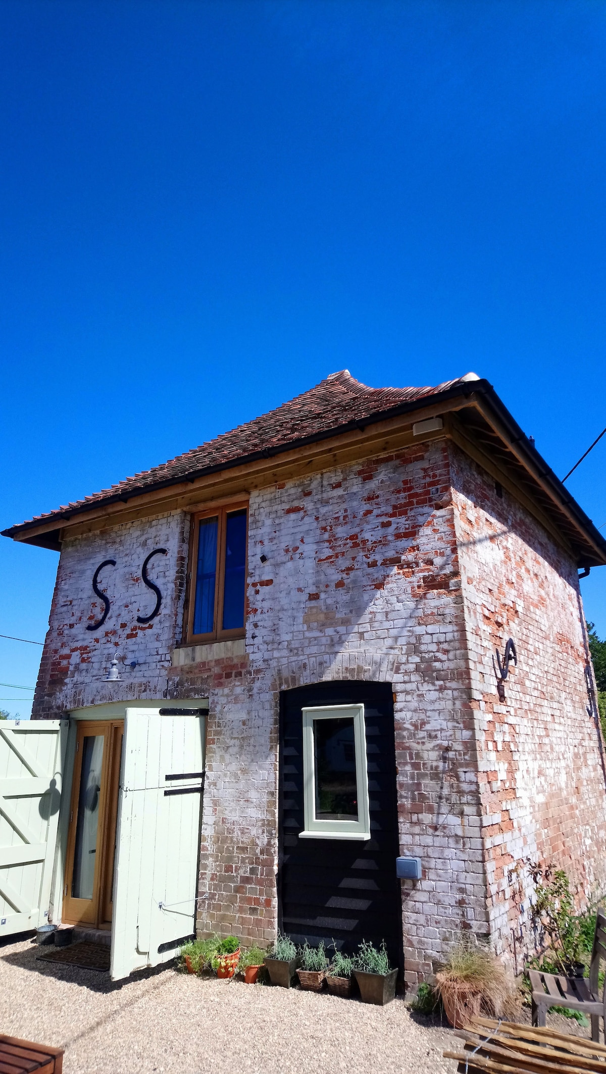 The exterior of The Dovecote is captured, highlighting the rustic brick façade with visible weathering. A small entrance is framed by the striking wooden door, flanked by a large window. The design features a charming pitched roof, with clear blue skies above.