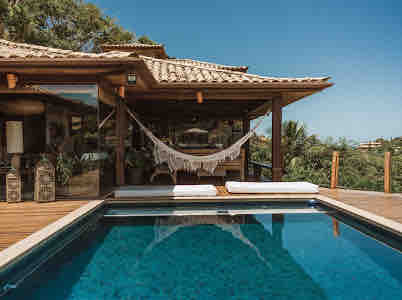 A private pool is framed by a wooden deck, featuring two sun loungers and a hammocking area. Tropical vegetation surrounds the space, with large windows visible in the background, reflecting a bright blue sky and providing a serene ambiance.