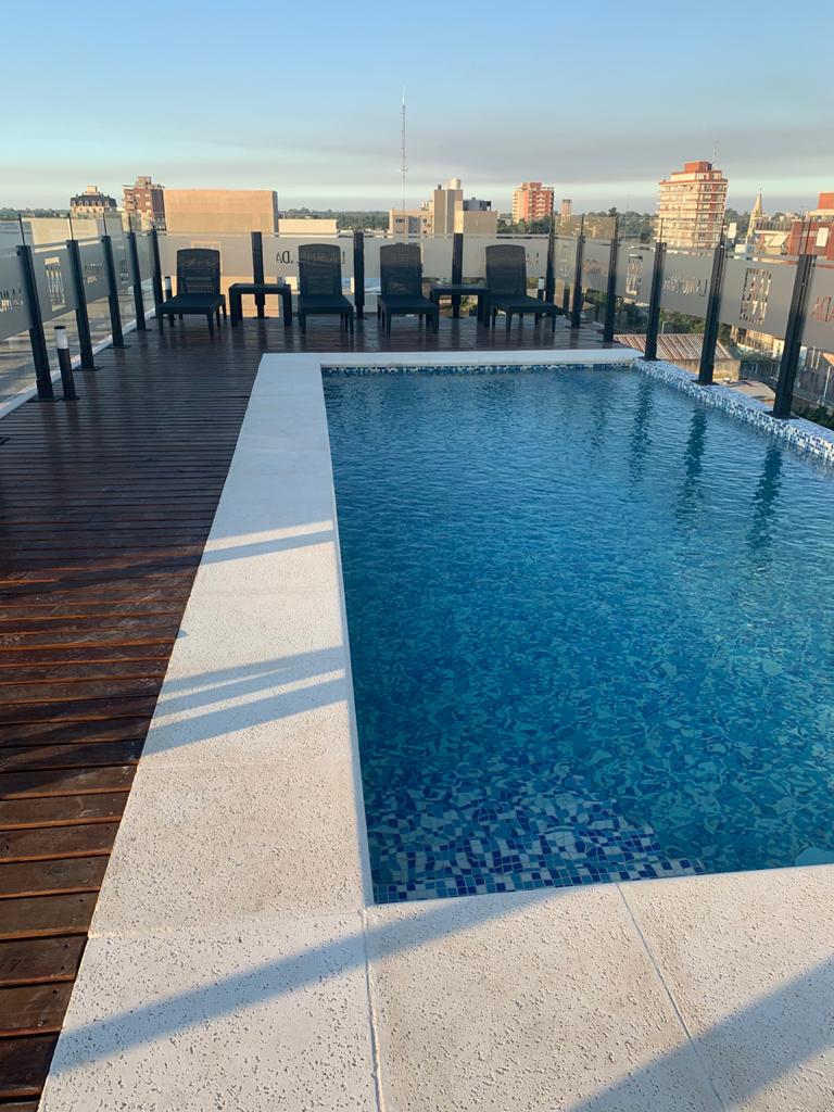 An outdoor terrace is shown with a swimming pool, its clear water reflecting the sky. Surrounding the pool are several black lounge chairs positioned on a wooden deck, providing an inviting space for relaxation. The city skyline is visible in the background.