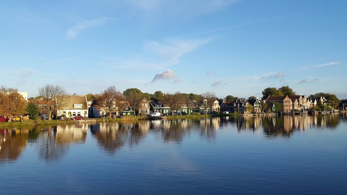 A serene view of a waterfront scene showcases a collection of charming houses lining the riverbank. The clear blue sky reflects in the calm water, creating a peaceful atmosphere. Surrounding trees add to the natural beauty of the setting.