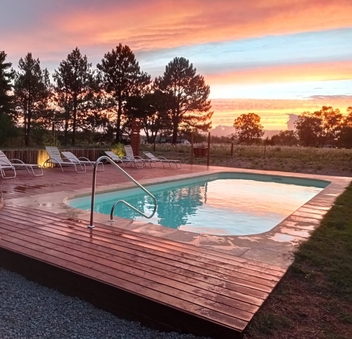 A private pool is set against a vibrant sunset, surrounded by wooden decking. Lounge chairs are arranged nearby for relaxation. Lush greenery and trees are visible in the background, enhancing the outdoor experience.