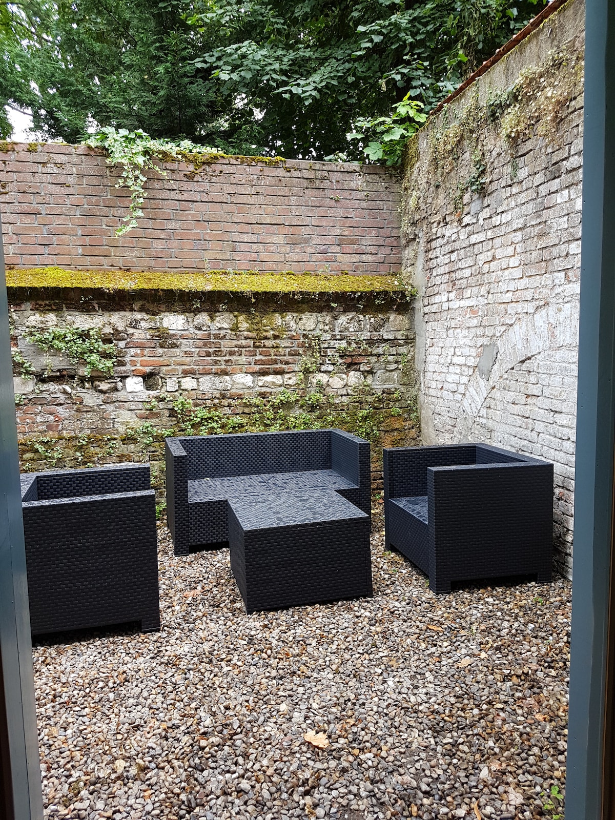 An enclosed courtyard is featured, showcasing a set of four dark wicker chairs surrounding a low table. Pebbles cover the ground, and a weathered brick wall provides a rustic backdrop, accentuated by greenery at the top.