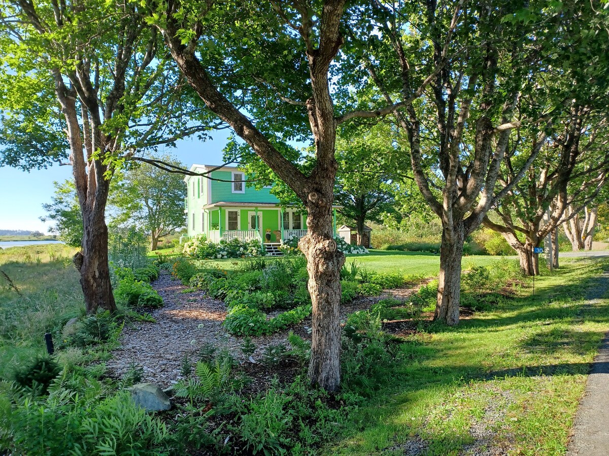 A charming green house is framed by mature trees and lush greenery. A gravel pathway leads to the entrance, providing a natural transition from the surrounding landscape. The view extends to a nearby waterway, enhancing the serene outdoor setting.