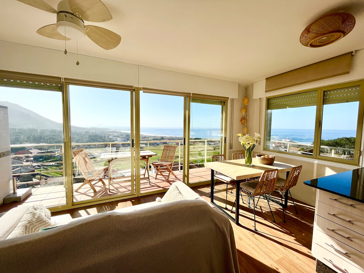 A bright living space features panoramic windows providing views of the mountains and the sea. A dining table for four is positioned near the windows, with comfortable chairs. Sunlight floods the room, highlighting the wooden floors and modern decor elements.
