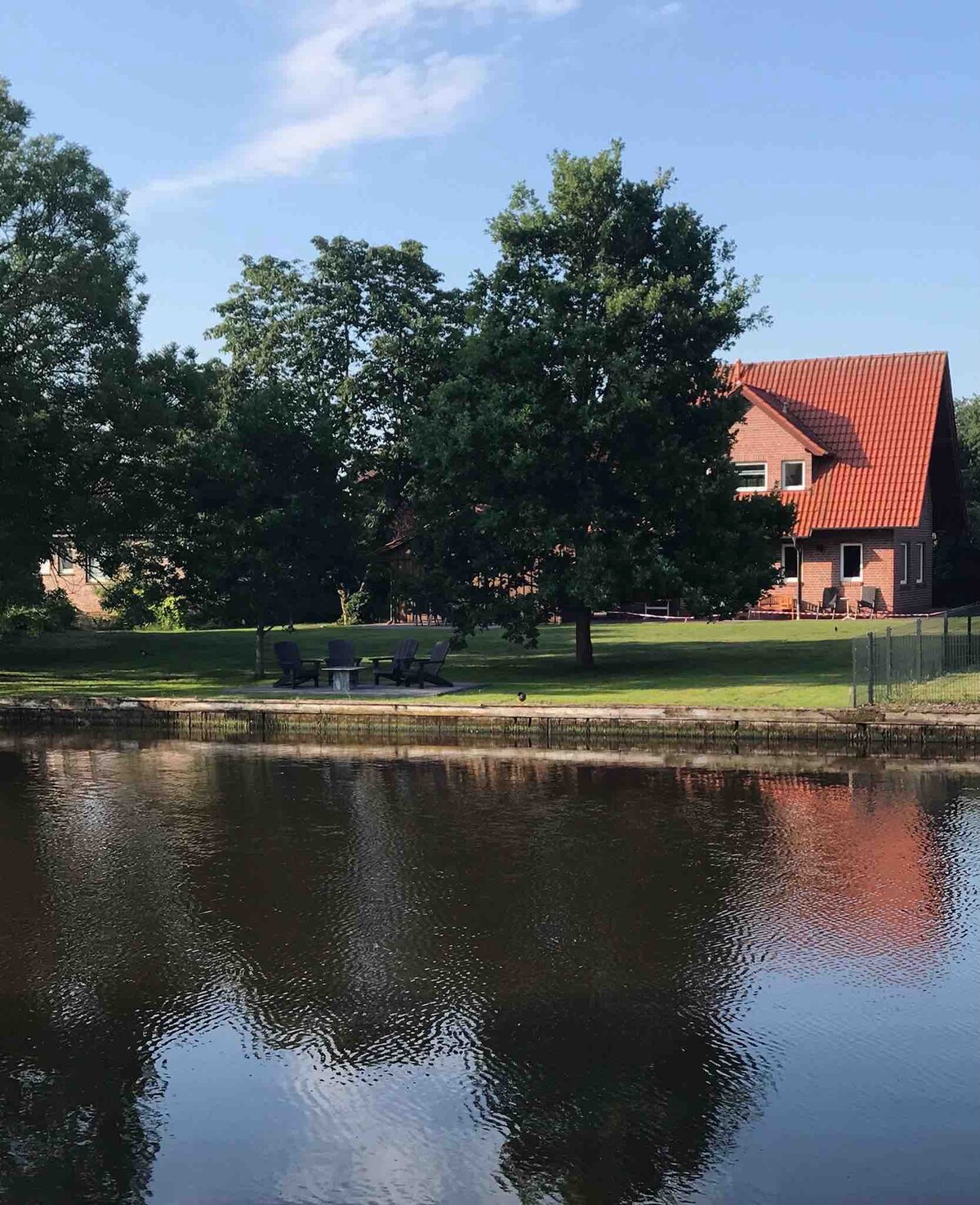 A well-maintained garden lies alongside a serene waterway, featuring a seating area with several outdoor chairs arranged under a large tree. A two-story brick house is positioned in the background, complemented by a red roof and surrounded by lush greenery.