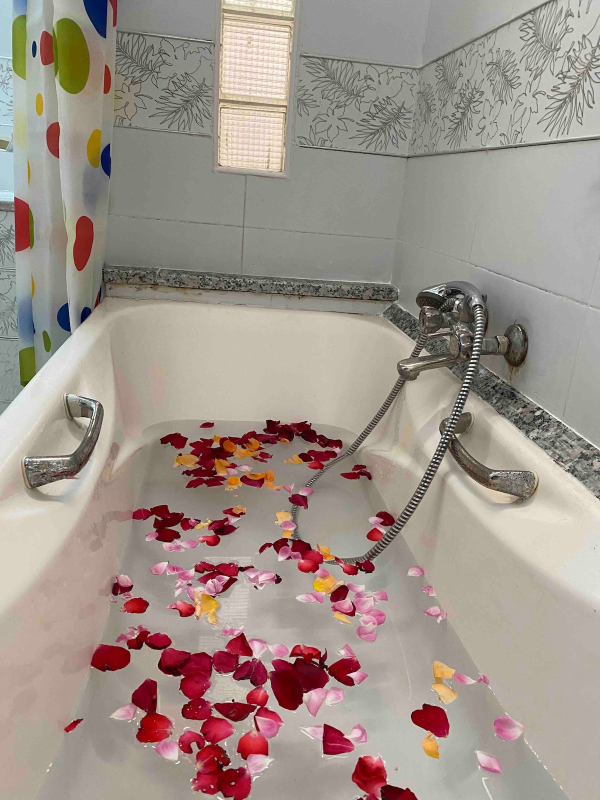 A clean bathtub is filled with water, adorned with scattered pink and yellow flower petals. The walls feature a decorative border with a floral motif, and a colorful shower curtain hangs nearby. Natural light enters through a small window.