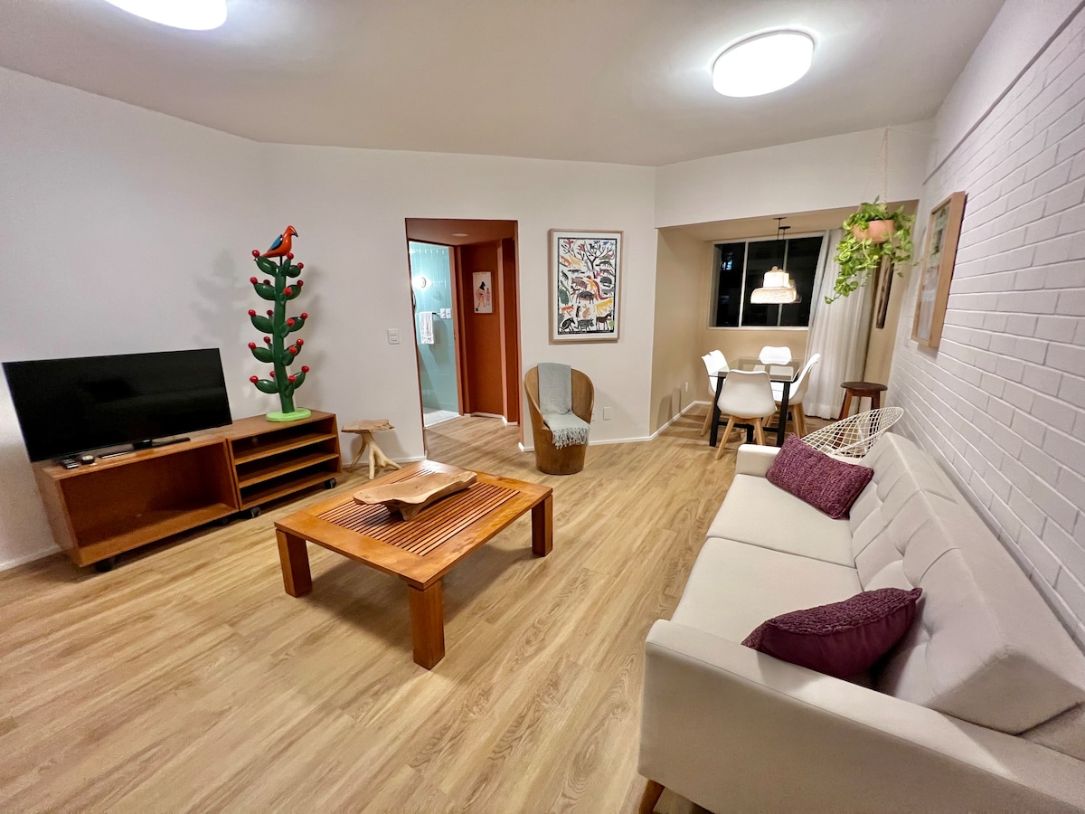 A spacious living area features a comfortable sofa with coordinating cushions, a wooden coffee table, and a TV on a media unit. A dining table with chairs is nearby, while decorative elements, including wall art and indoor plants, enhance the atmosphere.