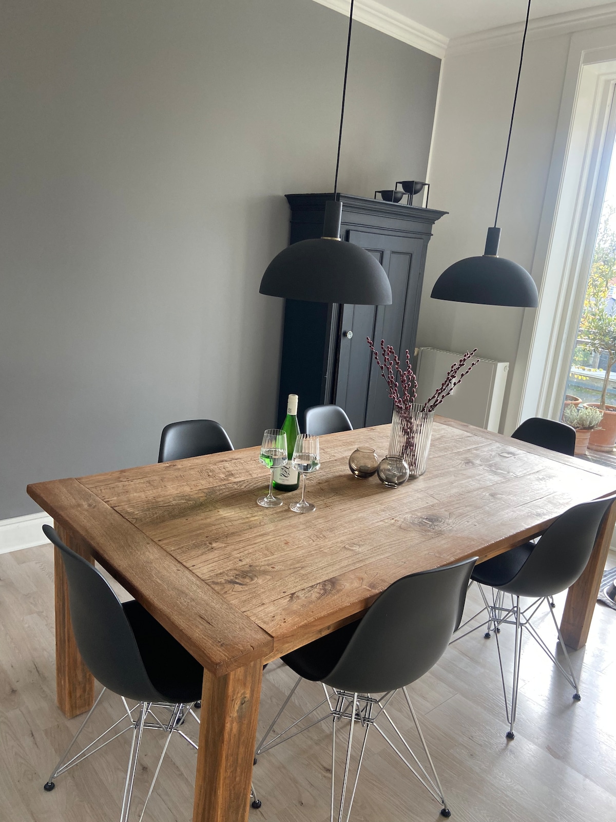 A wooden dining table is centrally positioned, accompanied by black chairs. Overhead, two black pendant lights provide soft illumination. A vase with decorative branches and two glasses are centered on the table, enhancing the inviting atmosphere. A dark cabinet is visible in the background.