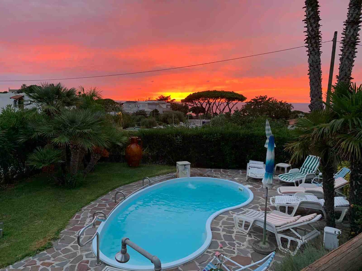A pool shaped like a heart lies centrally within a garden, surrounded by sun loungers and palm trees. The vibrant colors of a sunset fill the sky in shades of orange and pink, creating a serene atmosphere around the outdoor space.