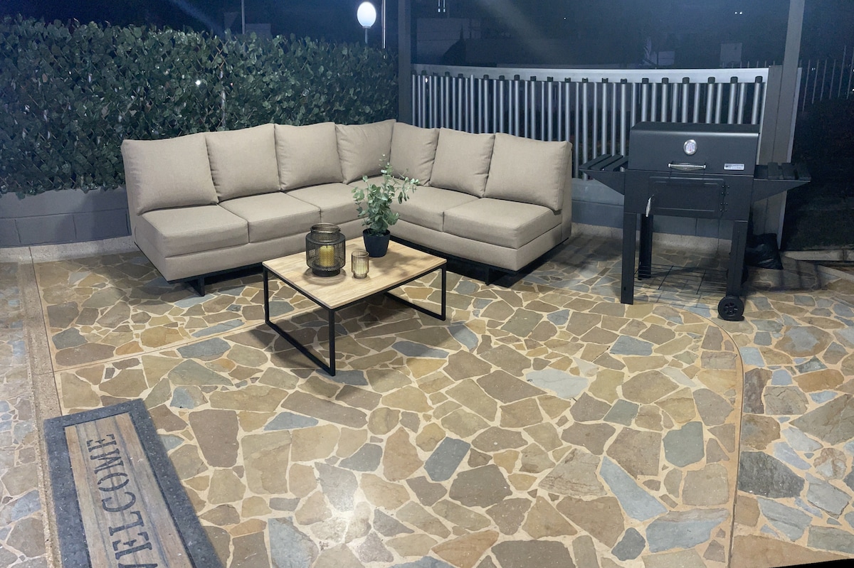 An outdoor patio area features a spacious sectional sofa in a neutral tone, complemented by a stone coffee table. A grill stands nearby, and natural stone flooring enhances the setting. A welcoming mat and green hedges provide an inviting backdrop.