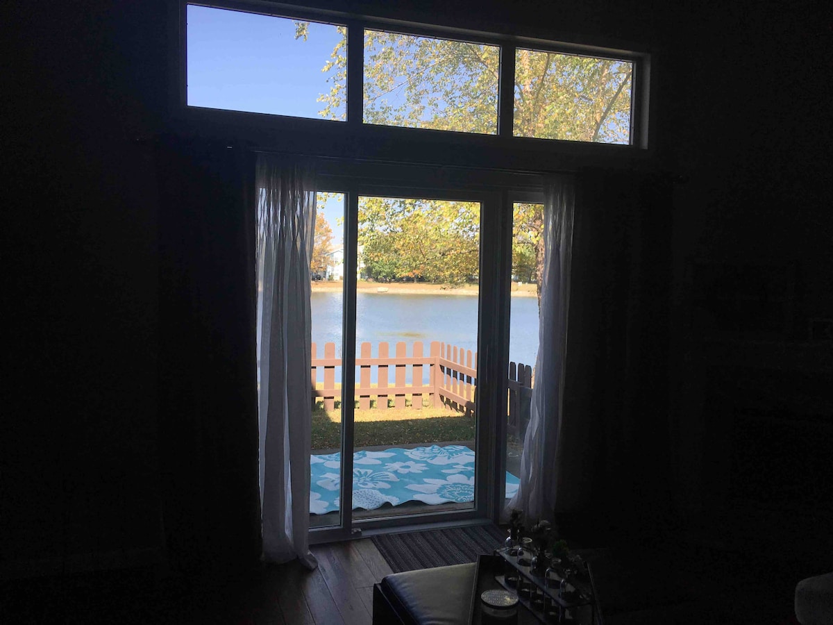 A view of Sandpiper Lake is framed by expansive sliding glass doors, allowing natural light to fill the room. Light sheer curtains gently sway, revealing a fenced outdoor space with a relaxing view of the water and nearby trees.