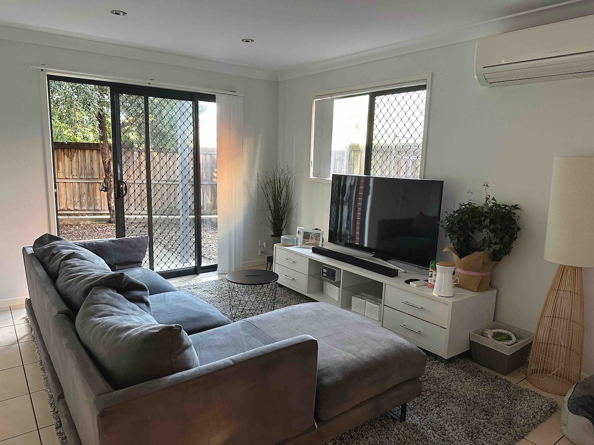 A comfortable living area features a large sectional sofa positioned near a wall-mounted television. Natural light enters through a large window and sliding glass doors overlooking a private outdoor space. A decorative plant sits beside the television unit, adding a touch of greenery.