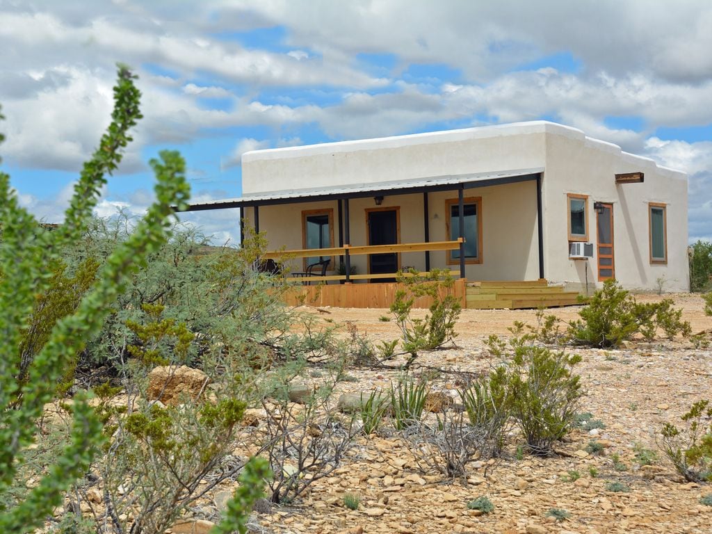 A single-story adobe home is set against a desert landscape, featuring a large covered porch at the entrance. The exterior walls are painted white, and the windows are framed in natural wood. Surrounding vegetation includes desert shrubs and small plants.