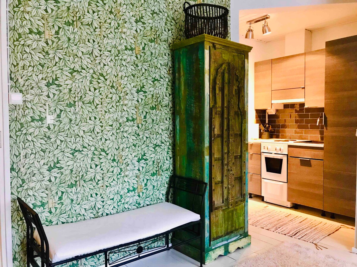A decorative corner features a bench with a white cushion against a leafy patterned wallpaper. An antique green cabinet stands nearby, adding a vintage element. The modern kitchen is visible in the background, with wooden cabinetry and appliances complementing the space.