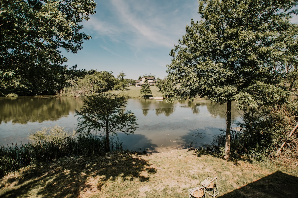 A serene view of a calm pond is framed by lush trees and greenery. Two chairs are positioned on the grass, inviting relaxation. A gentle slope leads to the water's edge, with a soft blue sky above and subtle reflections on the water's surface.