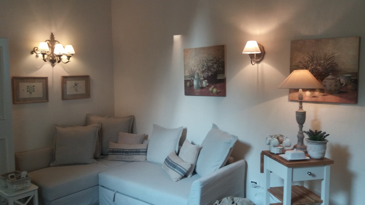 A bright living area is presented with a corner sofa adorned in neutral tones, complemented by soft cushions. Two wall fixtures provide gentle lighting, enhancing artwork featuring floral arrangements. A small side table is visible, supporting a decorative lamp and other items.