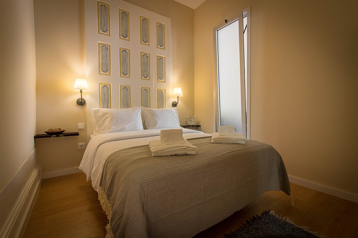 The bedroom features a spacious bed with a light-colored blanket and white pillows, flanked by wall sconces. Decorative wall art is displayed above the bed. A window allows natural light to enter, and minimalist furnishings contribute to a serene environment.