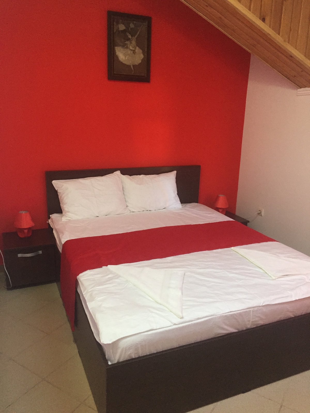 A bright room features a spacious bed with white linens and a red bedspread. Two bedside lamps provide soft illumination. A vibrant red wall serves as a focal point, while a piece of artwork is displayed above the bed. The floor is tiled, contributing to the room's clean aesthetic.