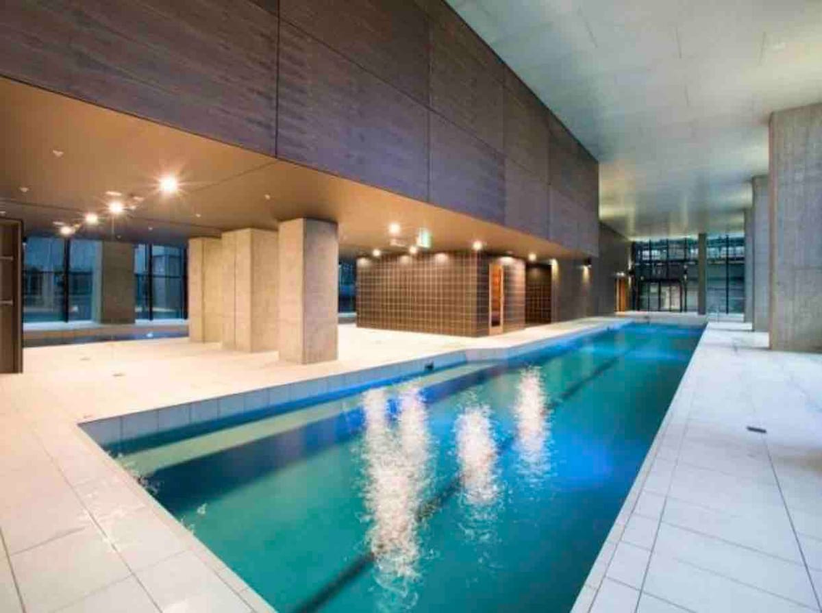 An indoor lap pool is presented, flanked by smooth stone pillars and sleek, modern walls. Ambient lighting creates a serene atmosphere, while large windows allow natural light to illuminate the space. The water reflects various shades of blue, inviting relaxation and tranquility.
