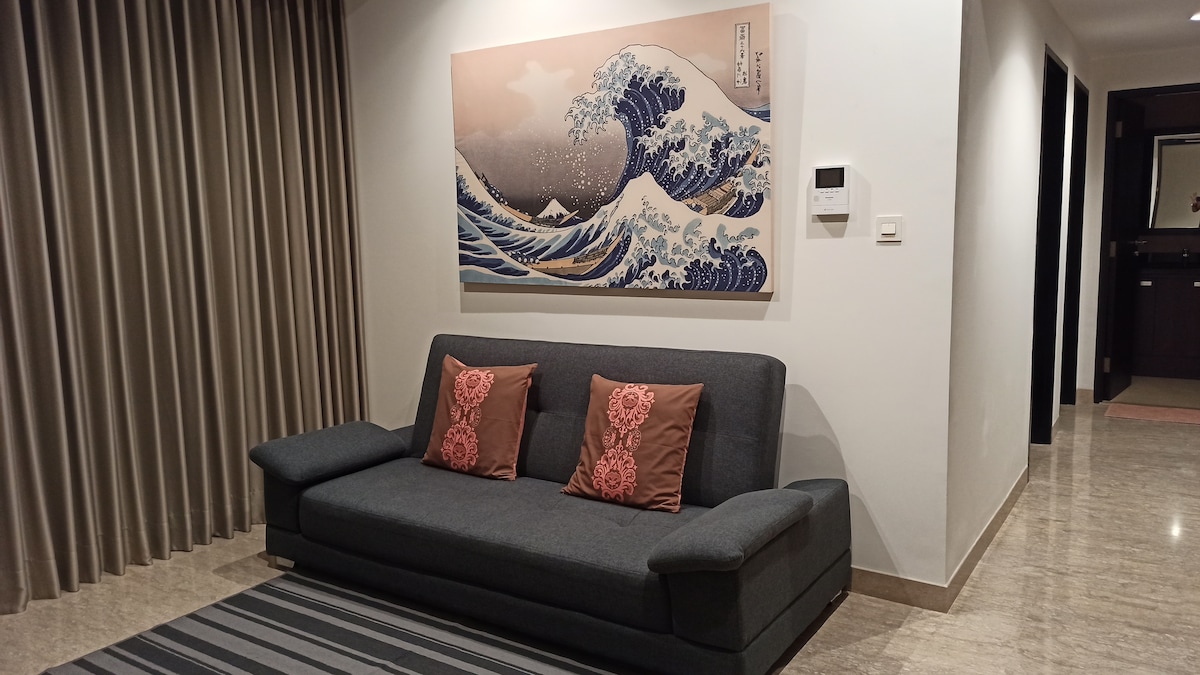 A modern living area features a dark grey sofa adorned with decorative pillows, positioned against a wall with a wave-themed artwork. Light beige curtains frame the window, while a striped area rug adds texture to the marble floor. The space conveys a clean, functional design.