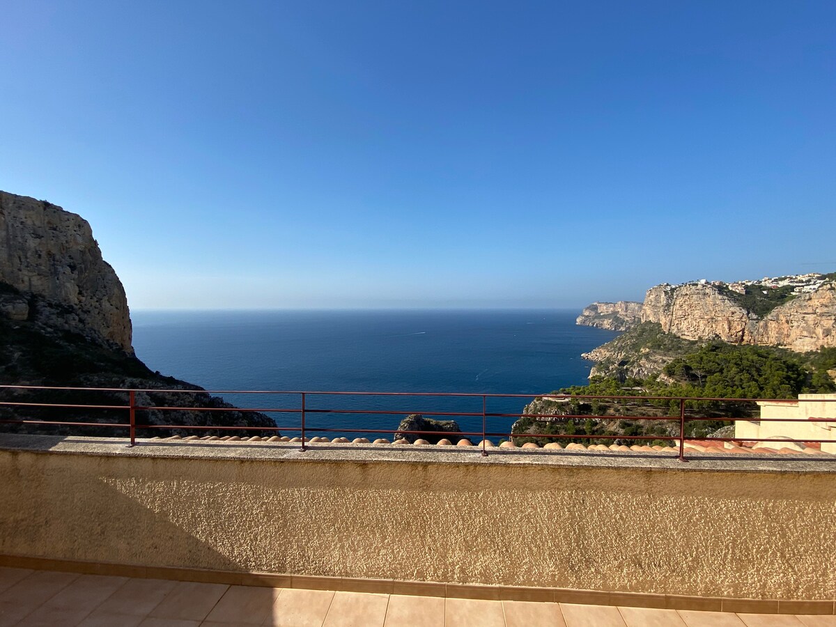 The spacious balcony offers a panoramic view of the Mediterranean Sea and surrounding cliffs. Clear skies create a serene atmosphere, highlighting the natural beauty of the coastline. The railing adds a touch of safety while allowing for unobstructed sightlines of the expansive ocean.