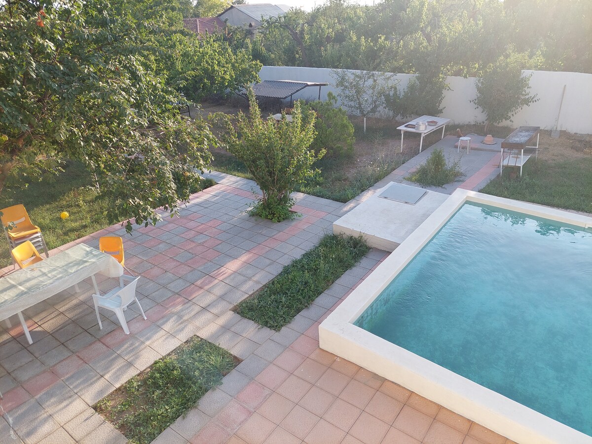 A spacious garden area features a refreshing swimming pool surrounded by well-maintained pathways and greenery. In the background, several outdoor seating areas are visible, including a table with chairs and recreational spaces for relaxation.