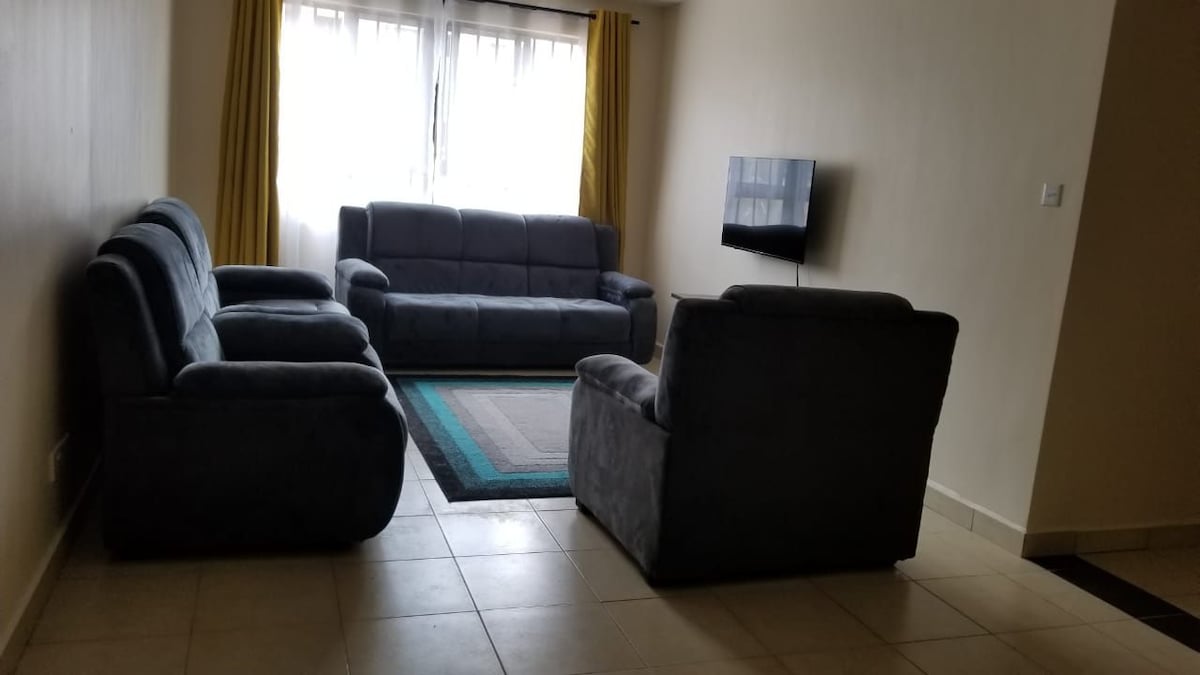 A comfortable living area features a set of soft, dark sofas arranged around a central rug. Large windows allow natural light to fill the space, with light curtains framing the view. A wall-mounted television is positioned opposite the seating area.