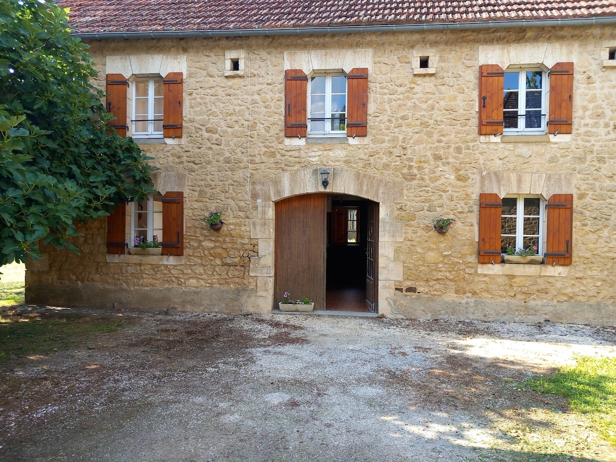 The exterior of a stone house is depicted, featuring wooden shutters and a large entrance adorned with a rustic archway. Flower pots are positioned near the windows, adding a touch of greenery. A gravel path leads up to the entrance, surrounded by a grassy area.