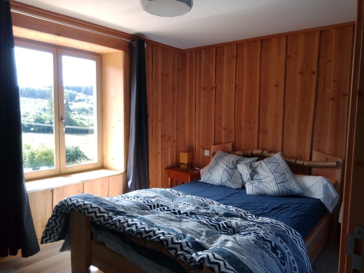 A warmly finished bedroom features wooden paneling and a spacious bed, dressed in a patterned blue quilt. Natural light filters through a large window, offering views of the surrounding landscape. Dark curtains frame the window, adding contrast to the room's light tones.
