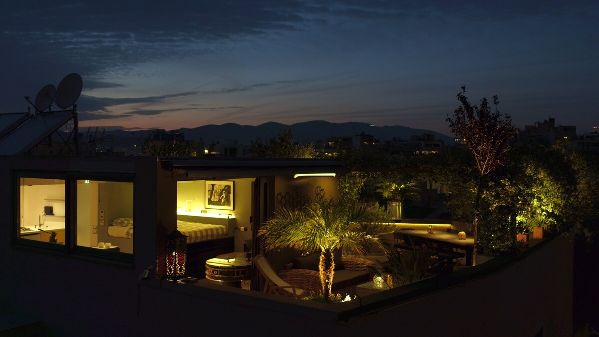 An evening view of a penthouse terrace, featuring warm lighting that enhances lush greenery and comfortable seating areas. In the background, soft silhouettes of distant mountains are visible against the twilight sky, while a cozy indoor space can be seen through large glass windows.
