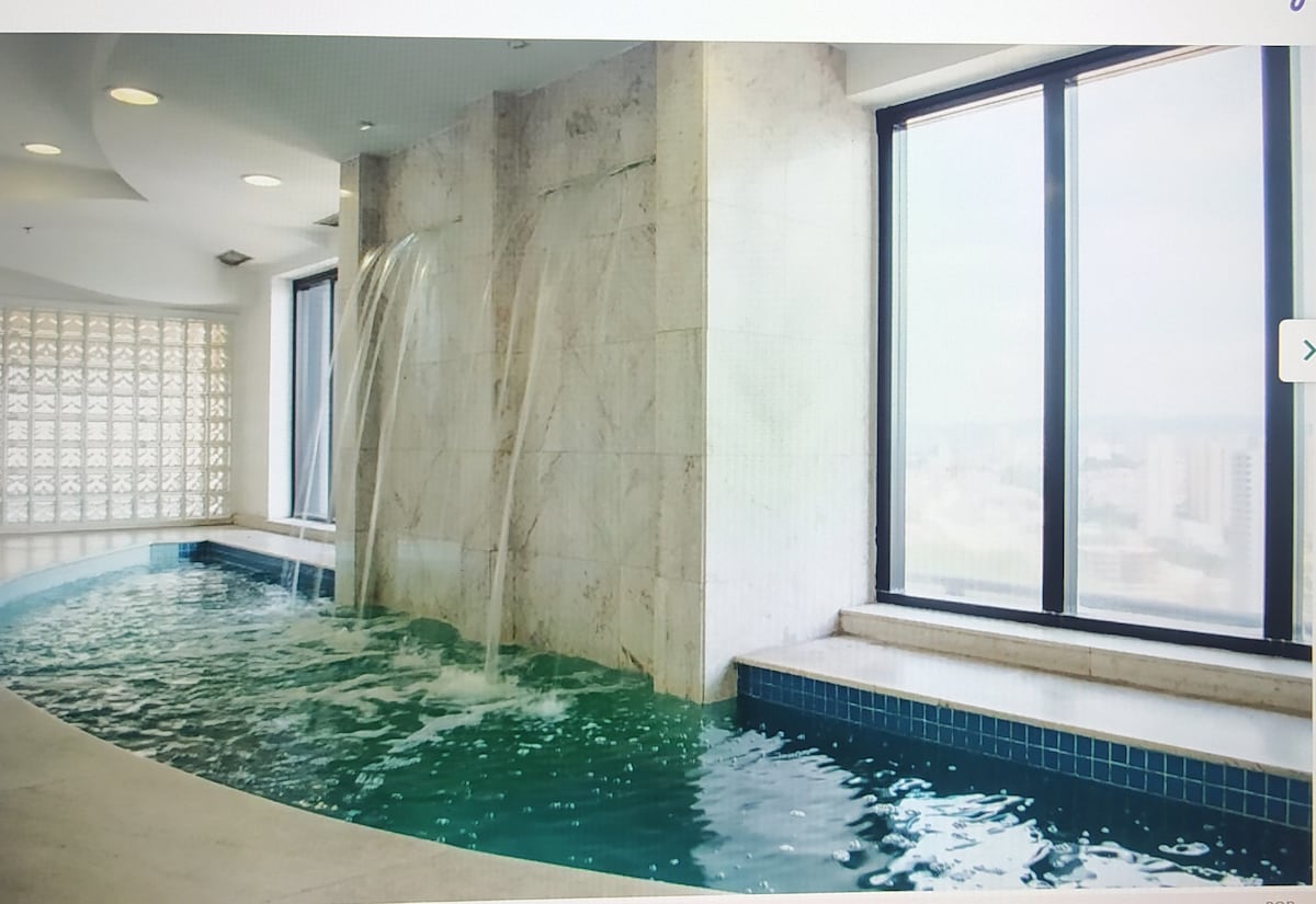 A tranquil indoor pool is captured with flowing water cascading over a stone wall. Large windows allow natural light to illuminate the space, creating a refreshing atmosphere. The pool is accented with blue tiles, inviting relaxation in a serene environment.