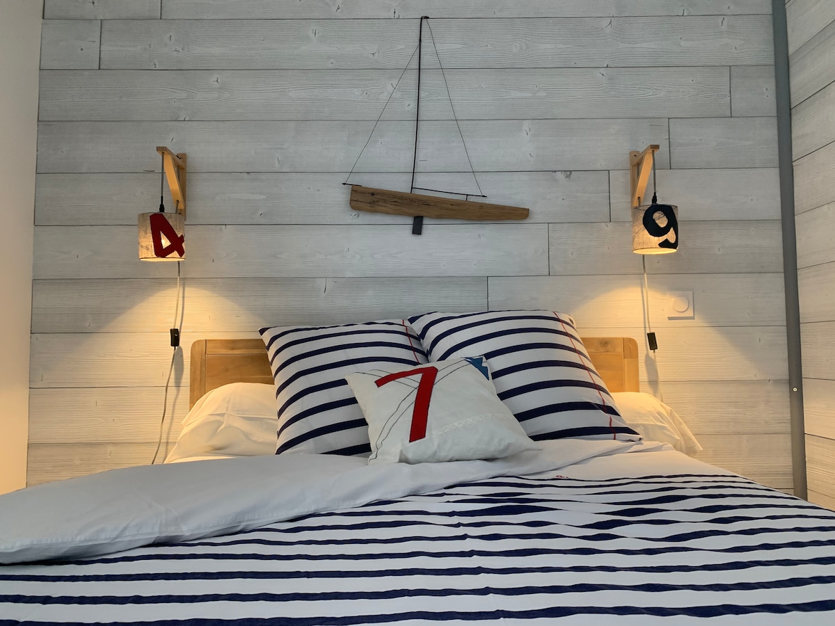 A cozy sleeping area features a spacious bed with navy and white striped linens. Two bedside lights illuminate the space, accompanied by decorative wall elements, including a wooden sailboat. Pillows in coordinating colors add a touch of comfort.