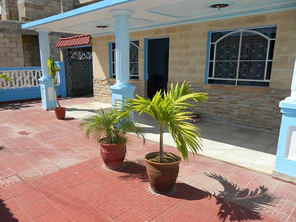 An outdoor area features a red tiled floor, framed by a low stone wall. Two potted palm plants add a touch of greenery near the entrance, which is adorned with columns and decorative stonework. A covered porch provides shaded seating space.