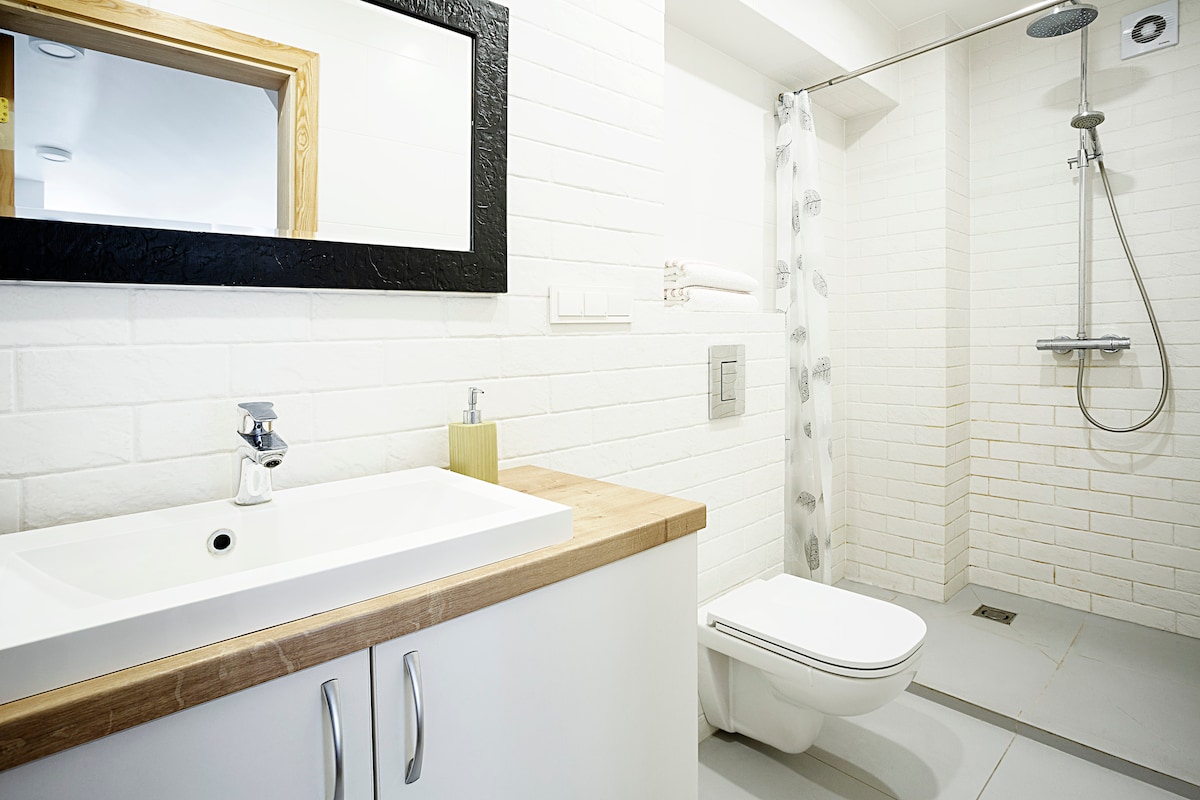 The bathroom features a modern design with white brick walls. A large mirror above the sink reflects a clean and functional layout. A rain shower is positioned next to the toilet, and a wooden countertop adds a touch of warmth to the space.
