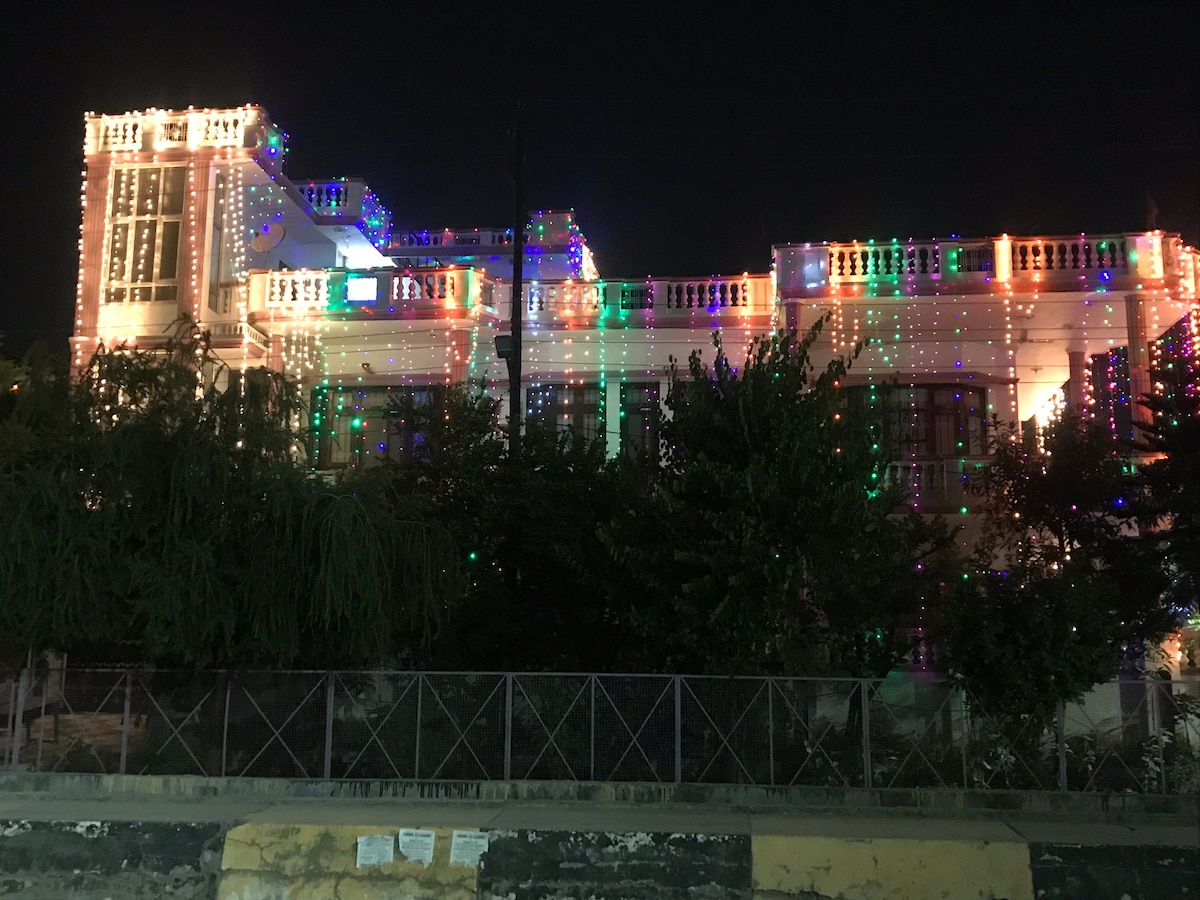 A well-lit building is adorned with colorful lights, creating a festive atmosphere. The structure features decorative railing and multiple levels. Nearby greenery is visible, contributing to the overall ambiance. The scene is set at night, highlighting the illuminated façade against the dark sky.