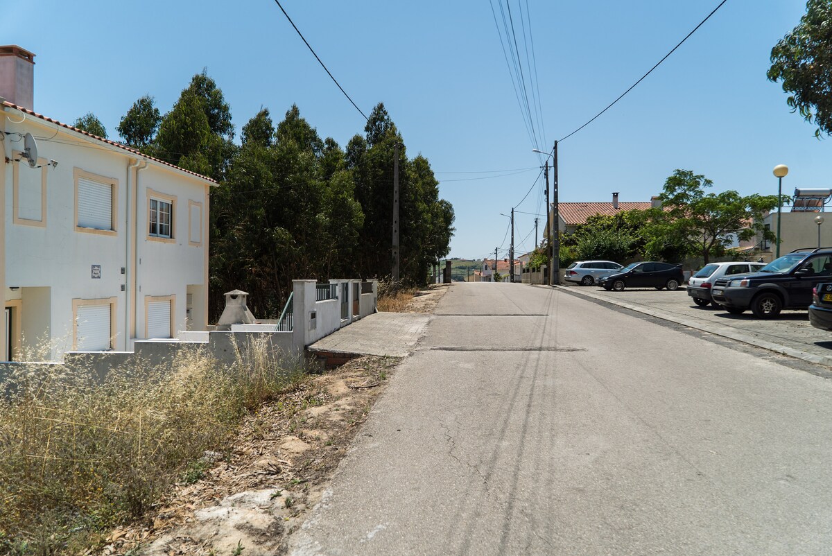 A quiet street is depicted, lined with trees on one side and residential buildings on the other. The road is paved and leads towards the horizon. Power lines extend along the street, providing a sense of accessibility, with nearby parking visible.