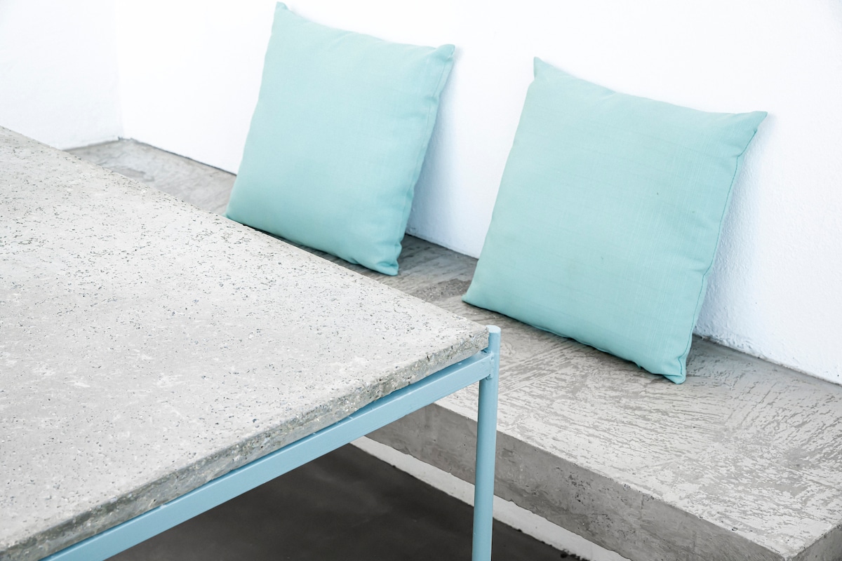 A modern dining area is displayed, featuring a minimalist concrete table paired with two soft turquoise cushions. The gentle color of the cushions contrasts with the neutral tones of the walls and floor, creating a calm and serene space.