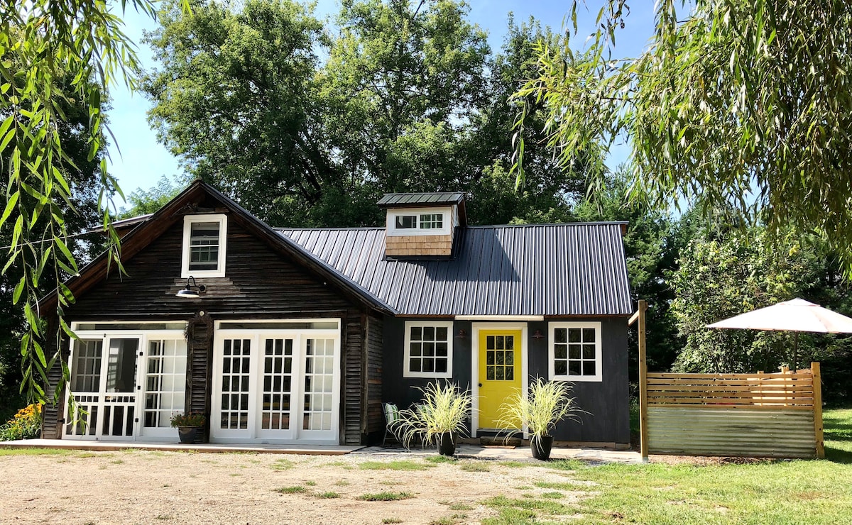 The exterior of the carriage house is showcased, featuring a combination of wood and modern elements. A bright yellow door contrasts with dark siding, and large glass doors invite natural light. Potted plants and a shaded area with an umbrella are visible in the outdoor space.