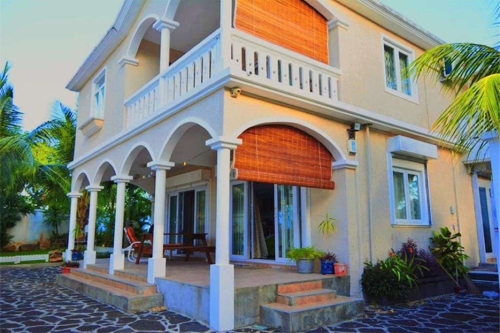 The villa exterior features a two-story structure with arched columns and wooden shades. A spacious terrace offers seating areas surrounded by tropical plants. The façade is light in color, complementing the stone pathway leading to the entrance, creating an inviting atmosphere.