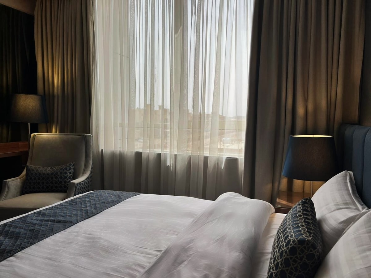 A well-appointed bedroom is featured, showcasing a spacious bed covered with crisp white linens. Soft light filters through sheer drapes, illuminating a cozy seating area with a plush chair and a bedside lamp on a wooden table.
