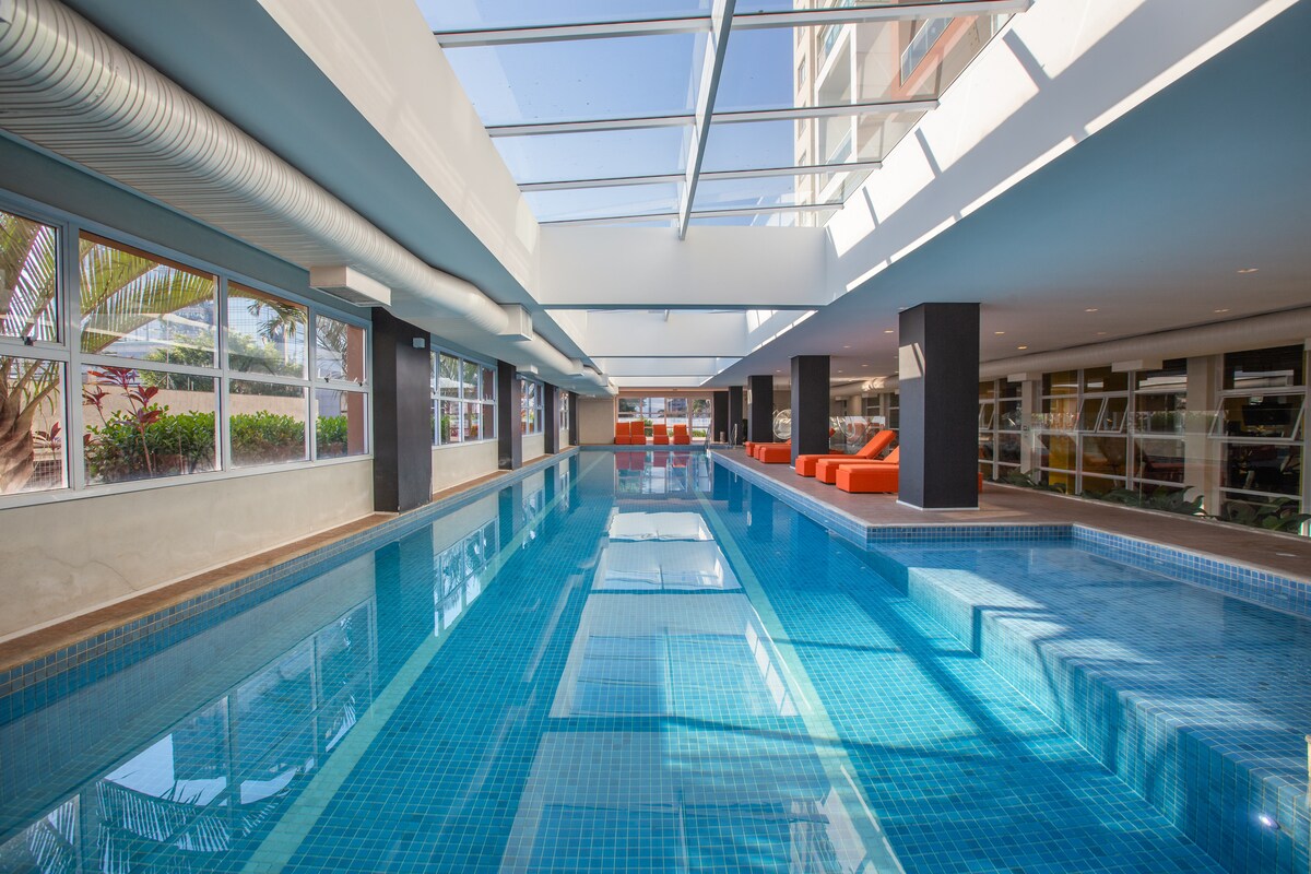 An indoor pool is displayed, featuring clear blue water and a spacious area surrounded by large windows that allow natural light to fill the space. Lounge chairs are positioned at the edge of the pool, offering relaxation options.