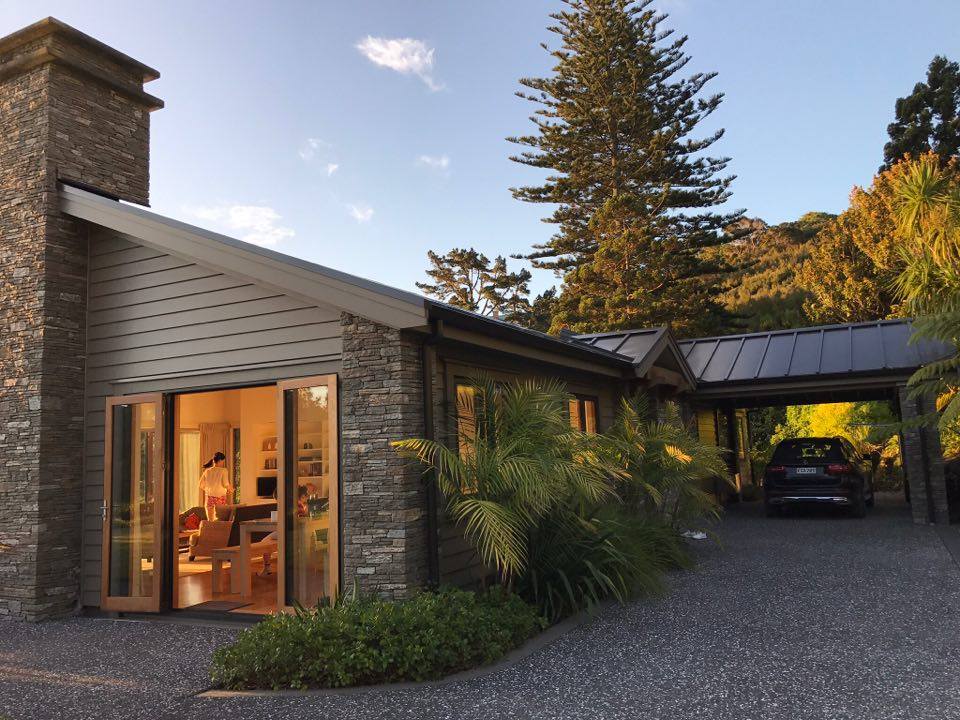 A stylish beach house entrance is framed by lush greenery and a gravel driveway. Large glass doors reveal a glimpse of the cozy living space inside, while a car is parked under an overhanging roof. Pine trees rise in the background, enhancing the serene setting.