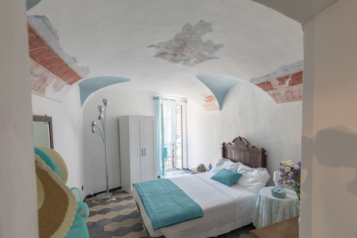 The spacious room features a combination of ancient frescoes and a modern design. A large bed is complemented by light blue accents, while natural light floods in through glass doors, leading to a small balcony. A wardrobe and a decorative table are also visible.