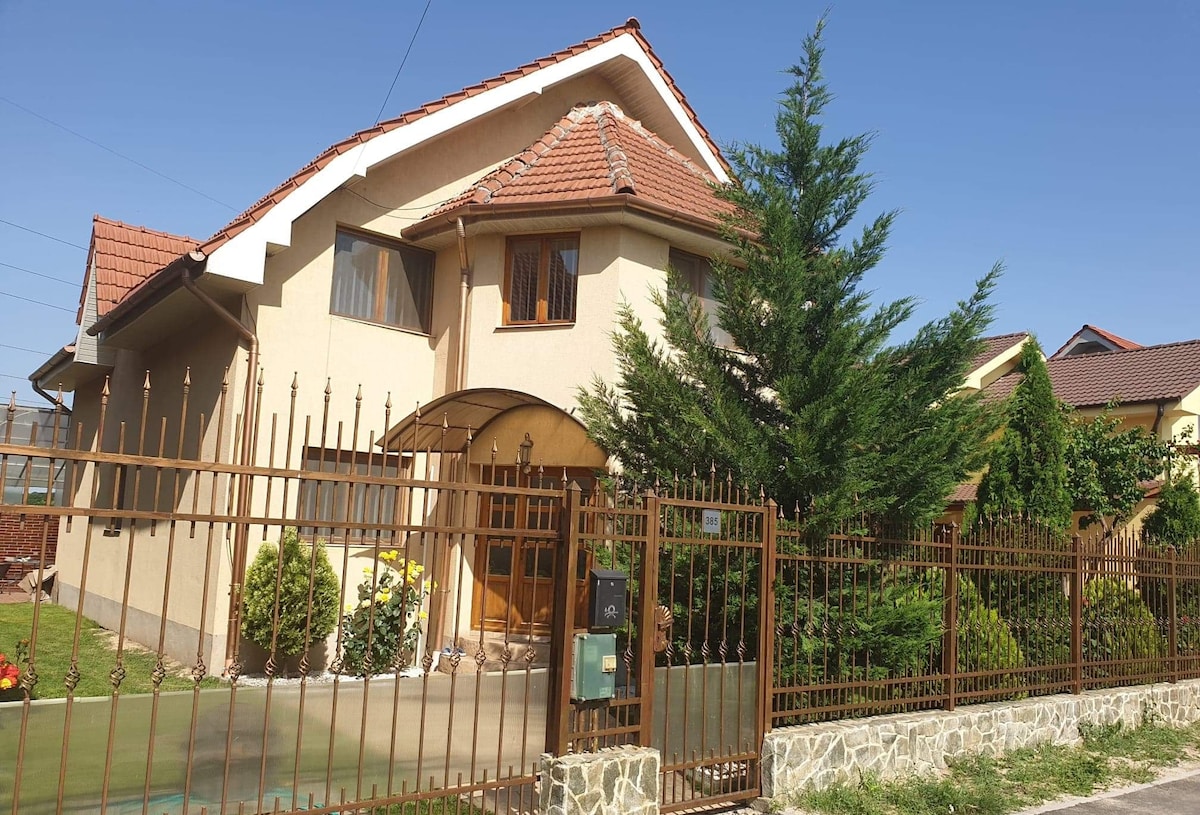 A two-story house is enclosed by a wrought-iron fence, showcasing a blend of beige and brown tones. Lush greenery and colorful flowers adorn the front yard, while the roof features a distinctive gable shape. Large windows invite natural light into the interior.
