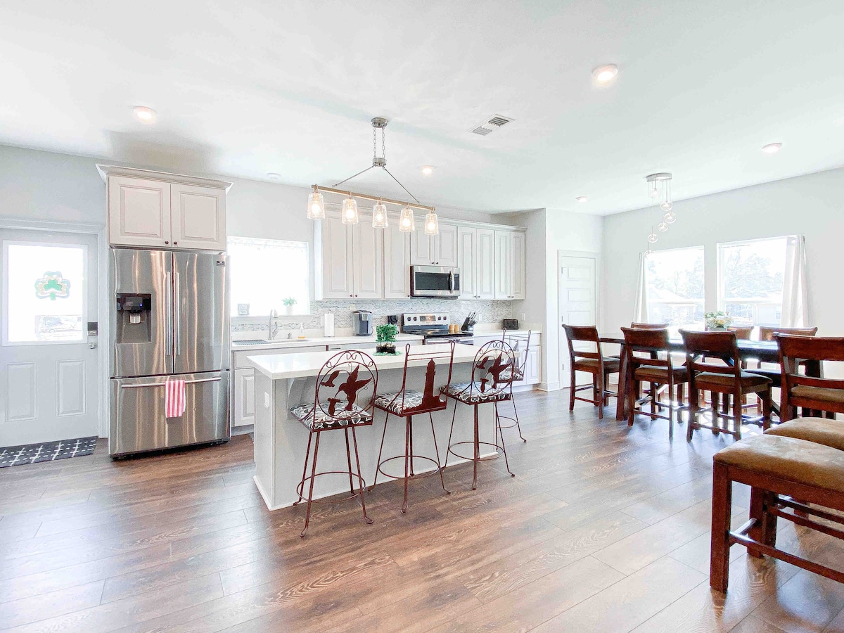 A modern kitchen is showcased, featuring stainless steel appliances and ample counter space. A central island with seating for four is surrounded by stylish bar stools. Natural light floods in through large windows, illuminating a spacious dining area that can accommodate several guests.