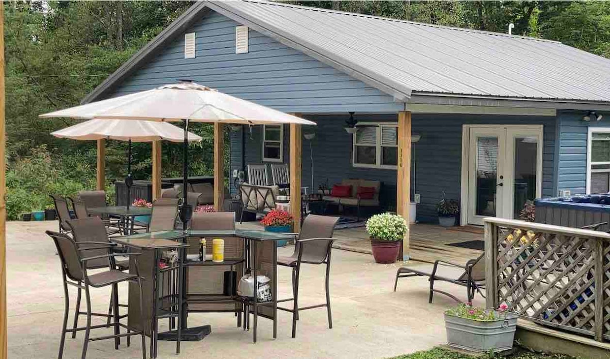 An outdoor seating area is furnished with multiple tables and chairs, shaded by a large umbrella. Surrounding the space are vibrant flower pots and a covered deck that extends from the home. The exterior features a blue facade and a modern metal roof.