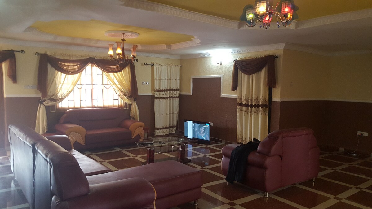 A spacious living area is furnished with large, comfortable sofas arranged around a glass coffee table. A television is visible, positioned against a wall. Natural light fills the space through a large window, complemented by decorative curtains.